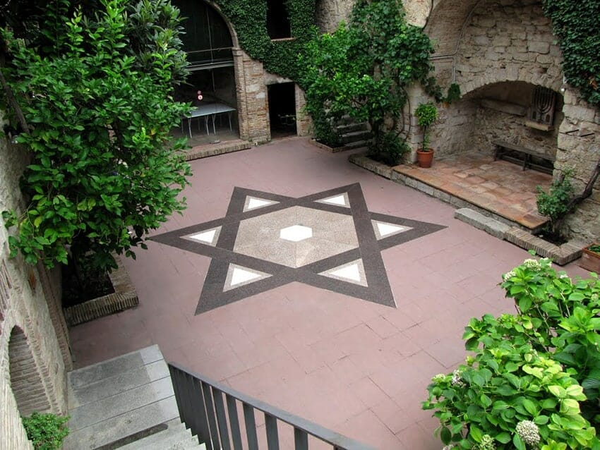Stone courtyard with a large black-and-white Star of David pattern in the center, surrounded by green plants, stone walls, and arched doorways.