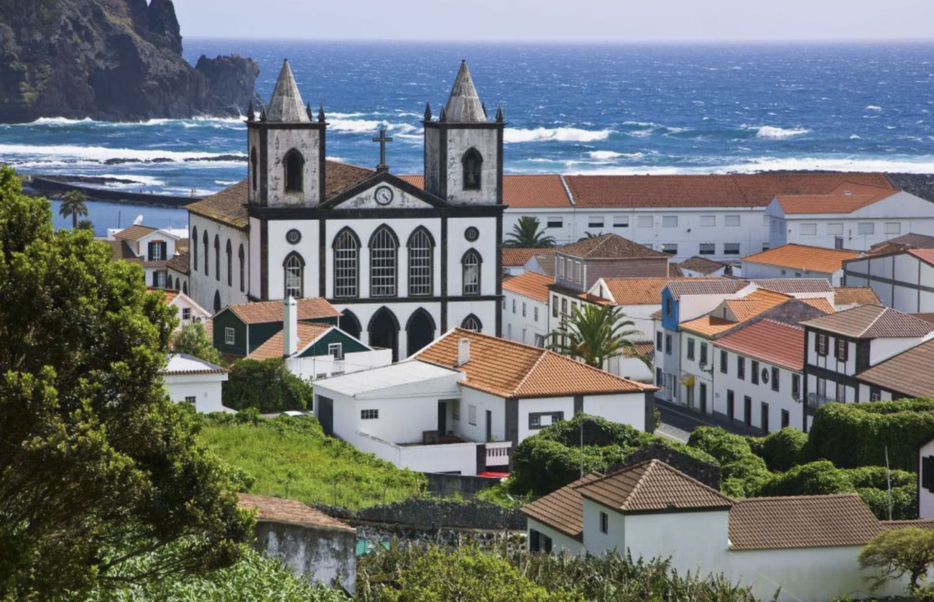 A white church with twin bell towers stands among red-roofed houses near the ocean, with waves and rocky cliffs in the background.