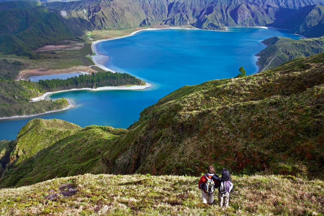 Two hikers stand on a grassy hillside overlooking a blue lake surrounded by green mountains and a forested peninsula.
