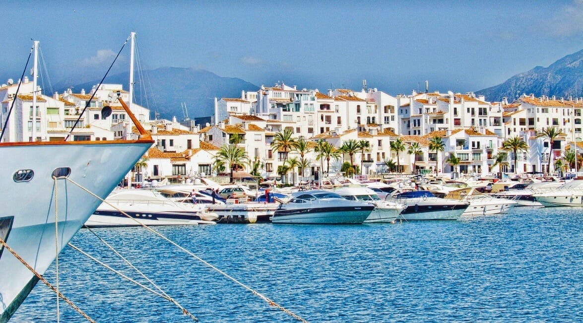 Boats and yachts docked at a marina with white buildings and palm trees in the background under a clear sky.