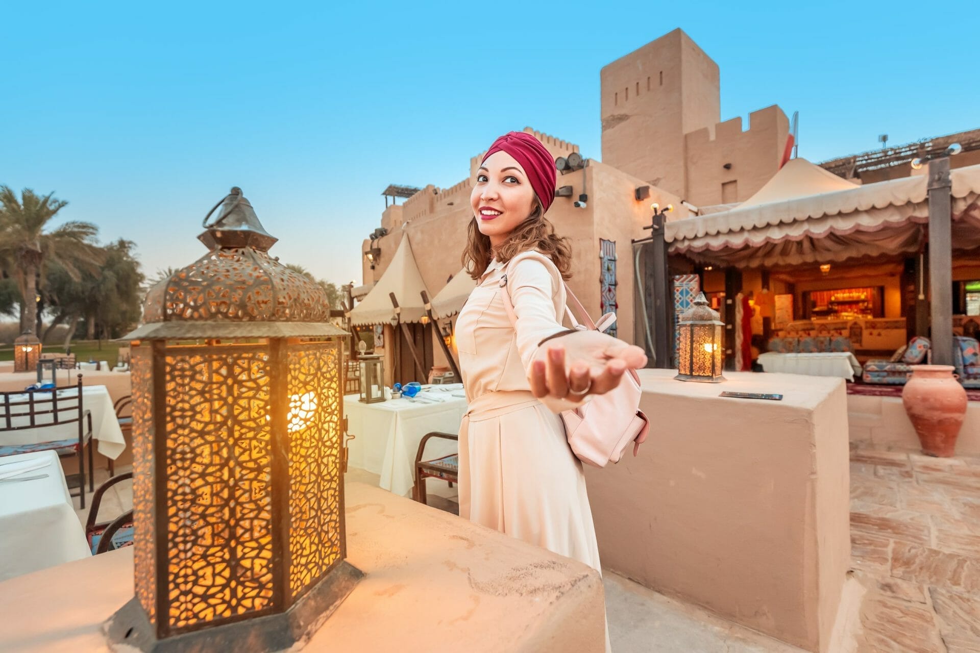 Morocco - woman in a white dress and red headscarf stands outdoors at a restaurant, holding out her hand and smiling, with a lit lantern in the foreground.