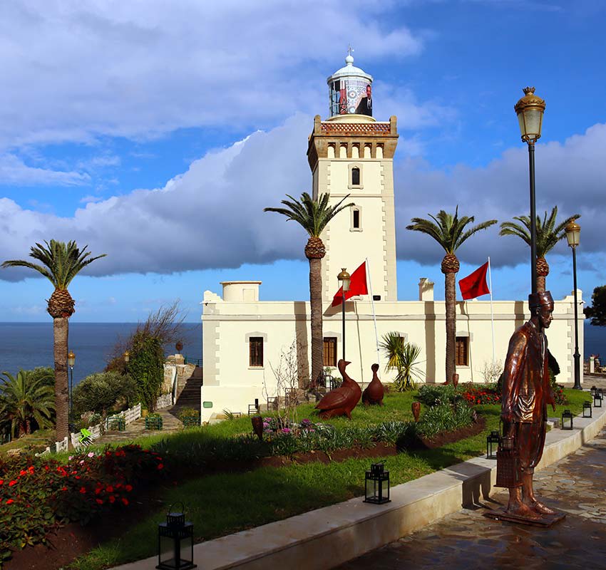 A lighthouse with a white tower stands by the sea, surrounded by palm trees, statues, two red flags, and garden landscaping under a partly cloudy sky.