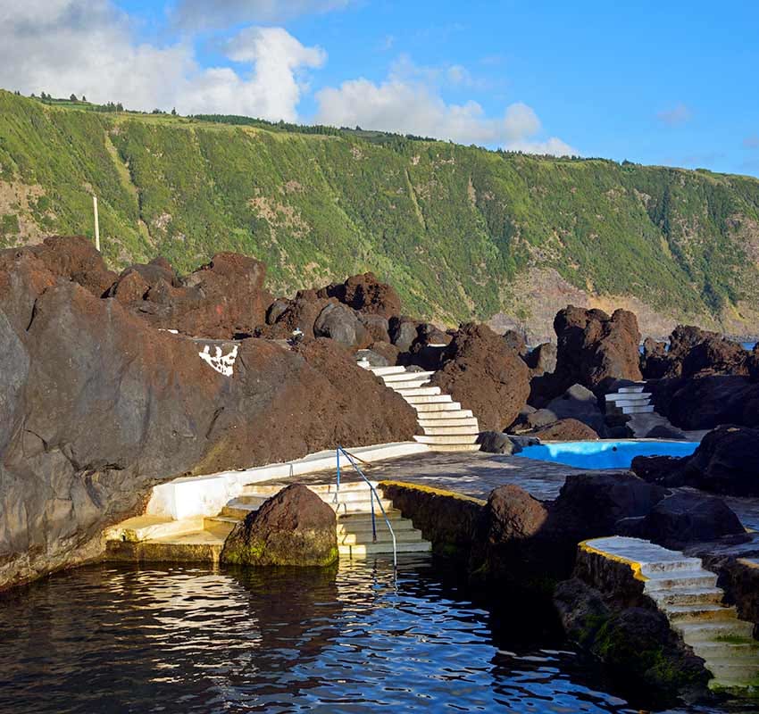 Concrete steps lead down to a natural swimming pool surrounded by dark volcanic rocks, with a green cliff and blue sky in the background.