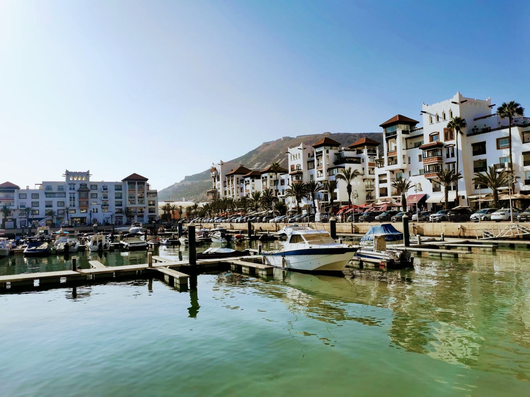 A marina with several docked boats, calm water, and multi-story white buildings with red roofs along the waterfront; a hill rises in the background under a clear blue sky.