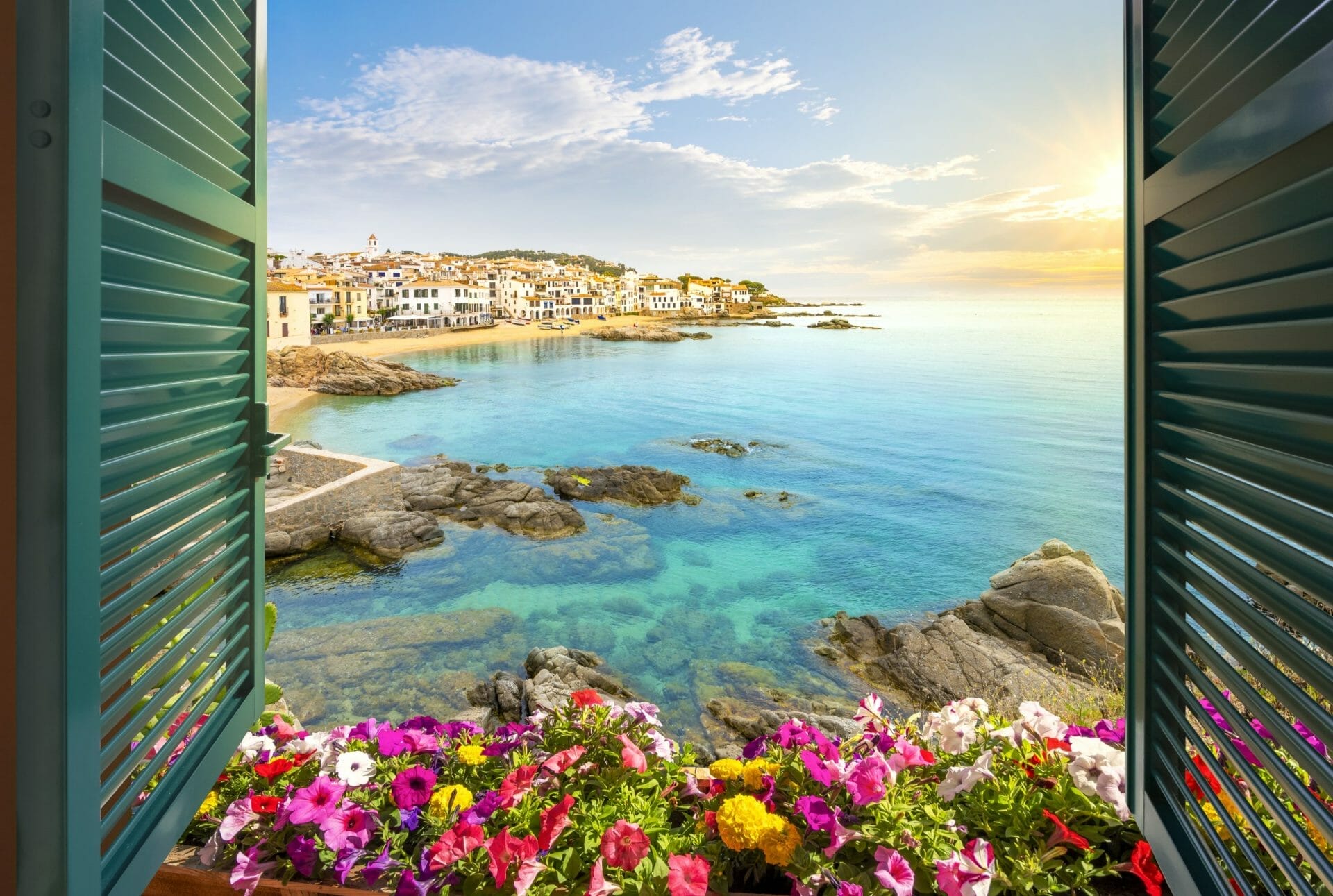 View through an open window with shutters of the sandy beach, rocky coastline and whitewashed town of calella de palafrugell, spain, on the costa brava coast as the sun sets.