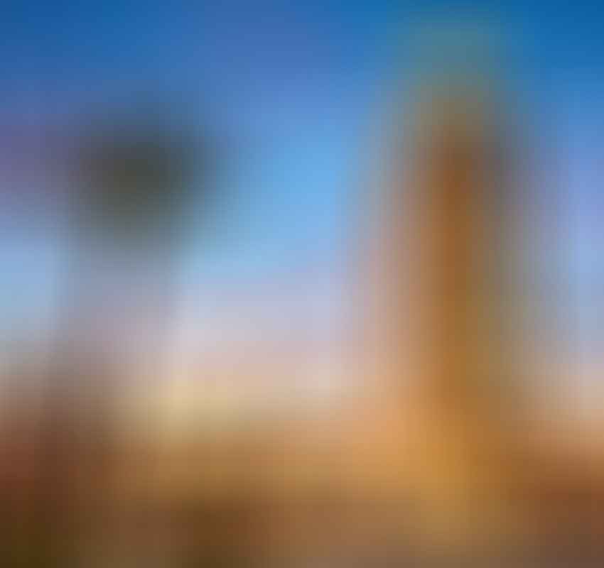 A tall stone minaret stands beside palm trees at sunset, with a colorful sky and illuminated lamps in the foreground.
