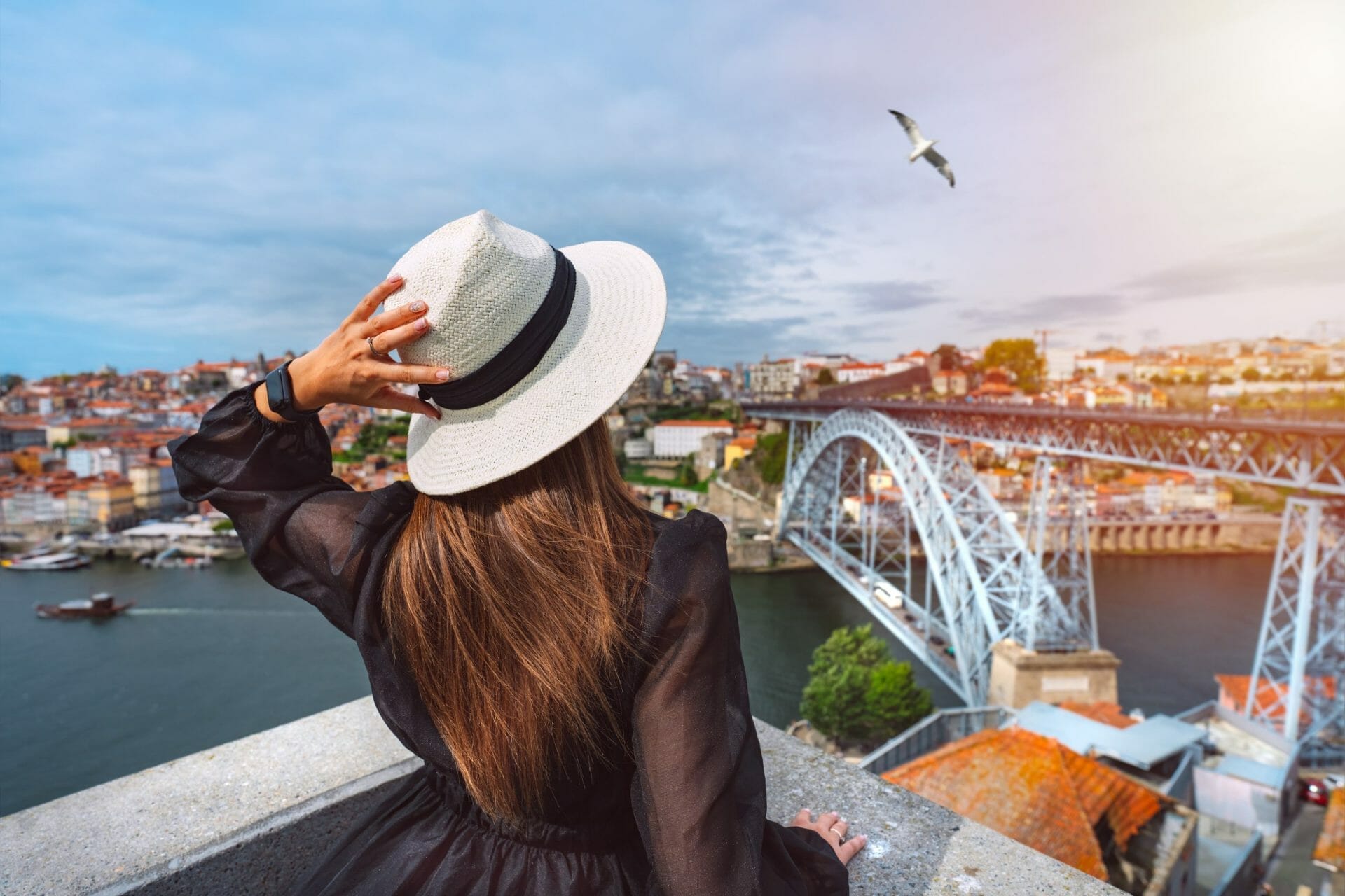A woman in a white hat overlooks a metal bridge and river, with a city in the background and a seagull flying above—an inspiring scene for Private Spain, Portugal or Morocco Luxury Travel adventures.