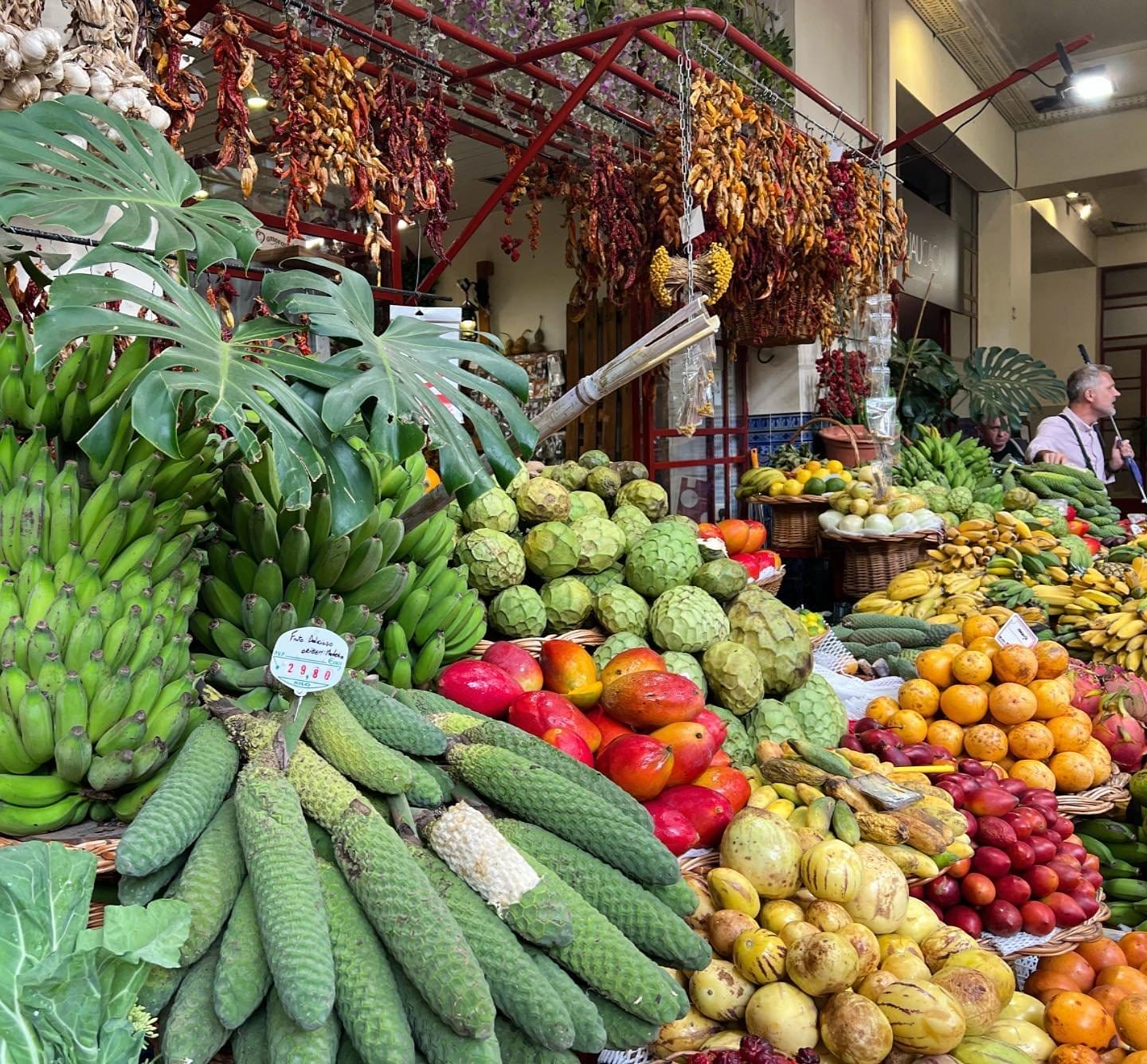 A Madeira Portugal market stall displays a variety of tropical fruits and vegetables, including bananas, pine cones, mangoes, and cherimoyas, with dried chilies hanging above—a vibrant scene often enjoyed on a Romantic Portugal Couples Private Luxury Tour.