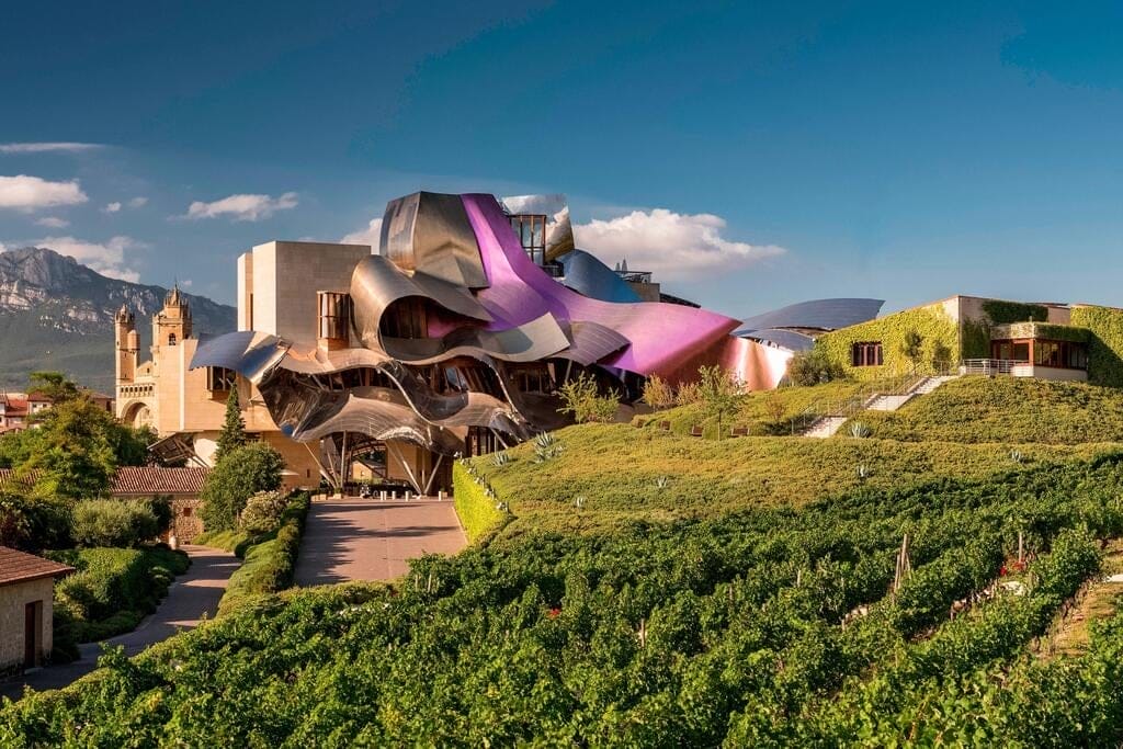 A modern building with curved metallic and colorful surfaces stands next to vineyards and traditional stone structures under a clear blue sky, offering a glimpse of the unique elegance found on Spain Portugal Morocco Private Luxury Tours.