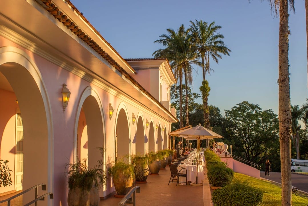 luxury spain portugal Outdoor dining area beside a pink building with arches, potted plants, and palm trees, set in warm sunlight.