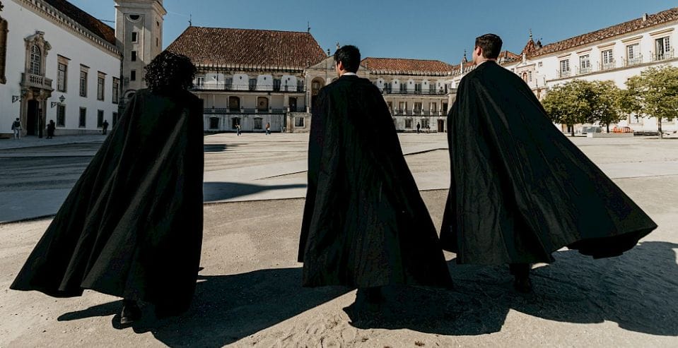 Three students in traditional black capes walking across the Pátio das Escolas, University of Coimbra, Portugal