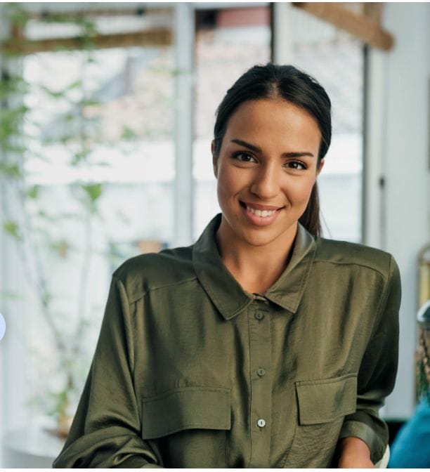 A woman with dark hair tied back, wearing an olive green button-up shirt, smiles while standing indoors near a window with plants in the background, ready to assist you as part of the Magical Private Travel Team.