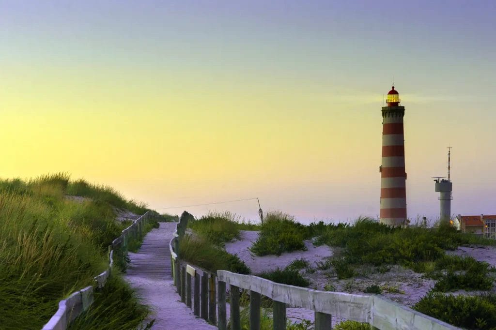 Tall striped lighthouse rising above the dunes at sunset on the Atlantic coast near Aveiro, Portugal