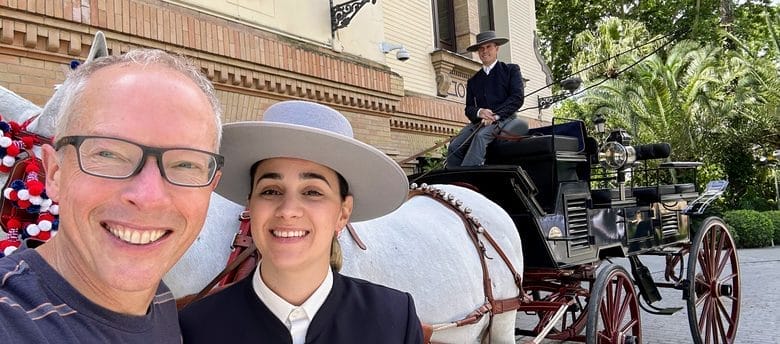 A man and woman in formal hats smile for a selfie in front of a horse-drawn carriage during their private luxury tour of Spain, Portugal, and Morocco.